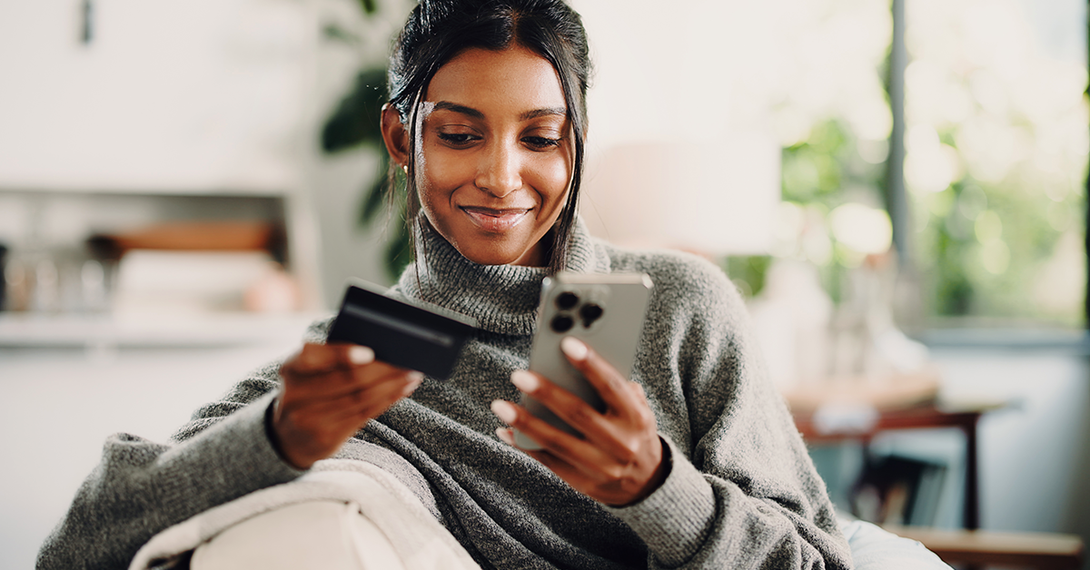 a smiling young woman looking at her credit card and phone while sitting on a couch