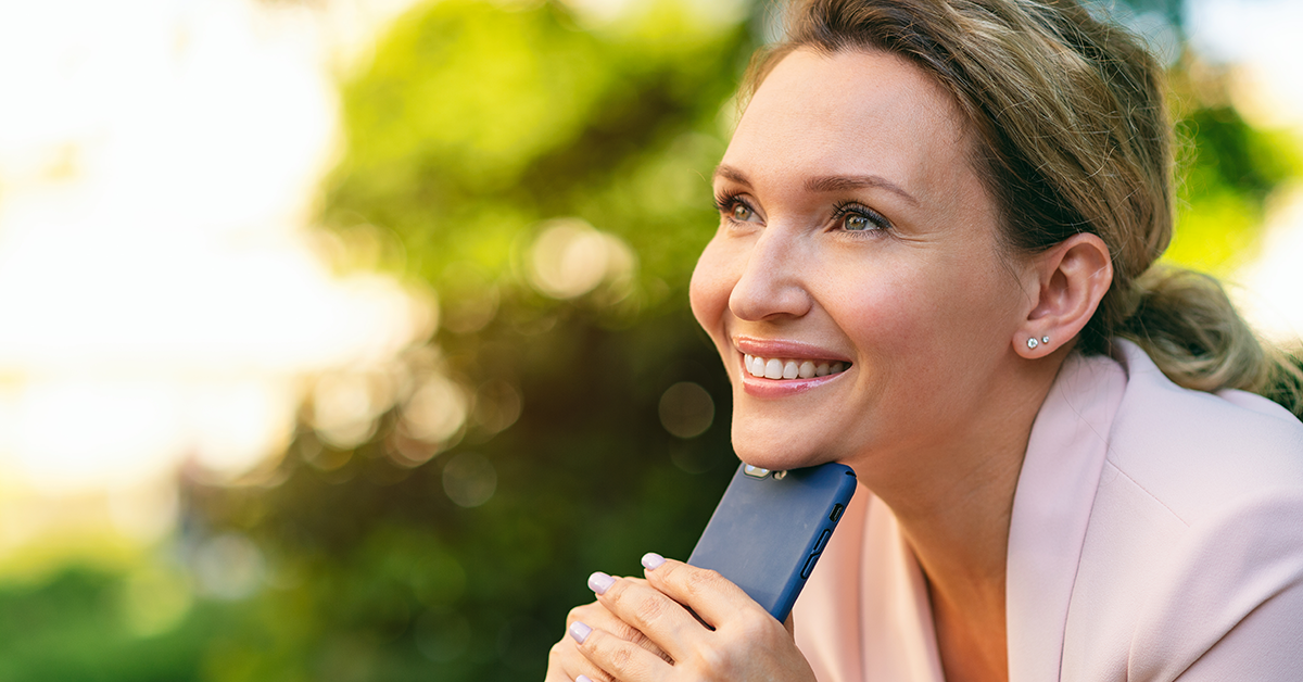 a woman holding her phone, smiling and looking thoughtful, while outside